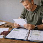 Älterer Mann prüft Dokumente, Reisepass und Versicherungsunterlagen am Tisch