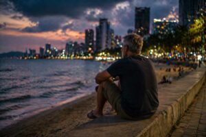 Älterer Mann sitzt alleine am Strand mit Blick auf Stadt – Einsamkeit im Ausland